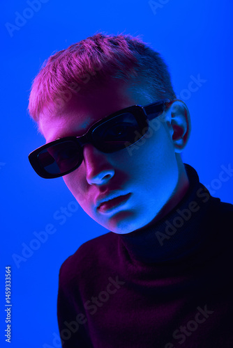 Obraz na płótnie Portrait of stylish young guy in sunglasses and black turtleneck posing against blue background in neon light. Close-up. Concept of bold fashion, style, youth culture, self-expression w salonie