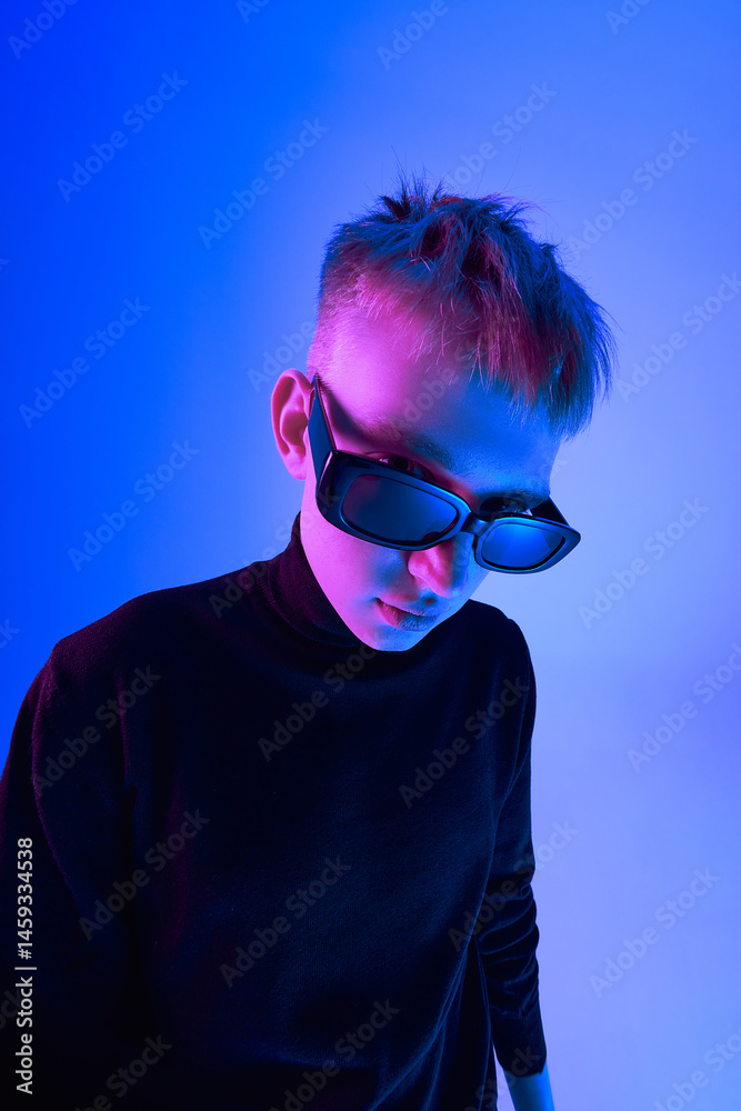 Close-up portrait of stylish young guy with short hairstyle, wearing sunglasses and black turtleneck, posing against blue background in neon light. Concept of bold fashion, style, youth culture