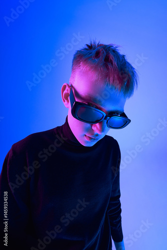 Obraz na płótnie Close-up portrait of stylish young guy with short hairstyle, wearing sunglasses and black turtleneck, posing against blue background in neon light. Concept of bold fashion, style, youth culture w salonie