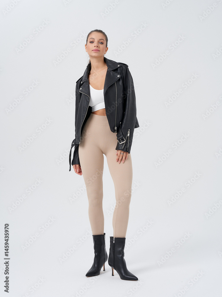 Fototapeta premium Woman with confident pose, wearing black leather jacket, white top, beige pants, and black heeled boots. Minimal makeup and hair in sleek bun, standing with hands by sides