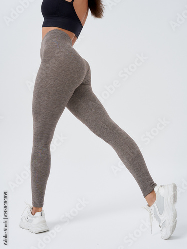 Athletic woman in dark sports top and gray leggings poses with one leg stretched out, wearing white sneakers. Photo captures part of her back, highlighting fitness and style