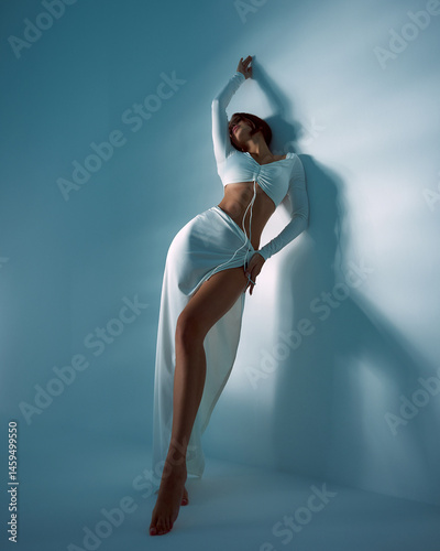 Tall model posing elegantly with an arched back, wearing a stylish white outfit with long sleeves and a high slit skirt, casting a soft shadow on the smooth surface