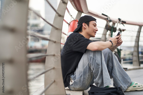 young Asian man sitting on ground chatting on smartphone at waterfront photo