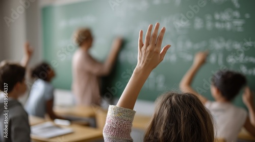 A closeup of a teachers hands writing mathematical equations on a chalkboard with eager students in the background raising their hands to participate.