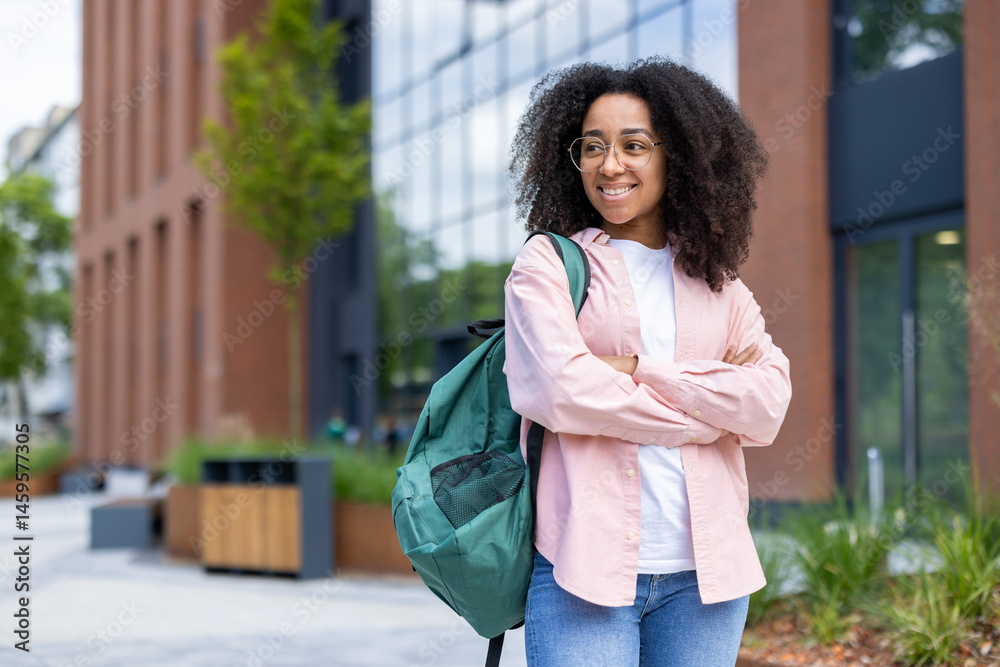 A young woman with a backpack stands confidently with arms crossed, smiling near a modern building on a sunny day. She is wearing glasses.