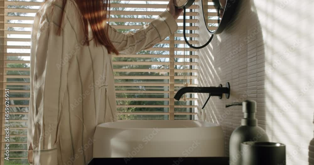 A woman gently opens wooden blinds in a modern bathroom, letting sunlight fill the room. The focus is on creating a tranquil ambiance and the beauty of simple living.