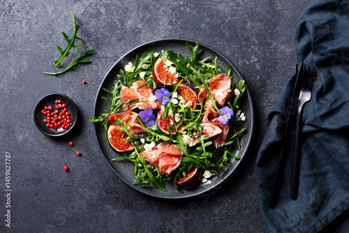 Arugula salad with fresh figs and prosciutto, jamon and edible flowers in a plate. Grey background. Top view.