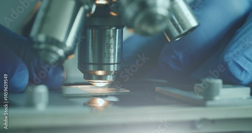 Close-up of biologist gloved hands using microscope in sterile laboratory. Vibrant blue gloves highlight precise scientific work against monochromatic lab backdrop