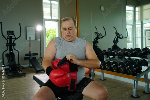 Elderly Caucasian man with a fat belly wearing gym clothes wearing red gloves practicing boxing and punching doing exercises for health and lose weight in a fitness centere.