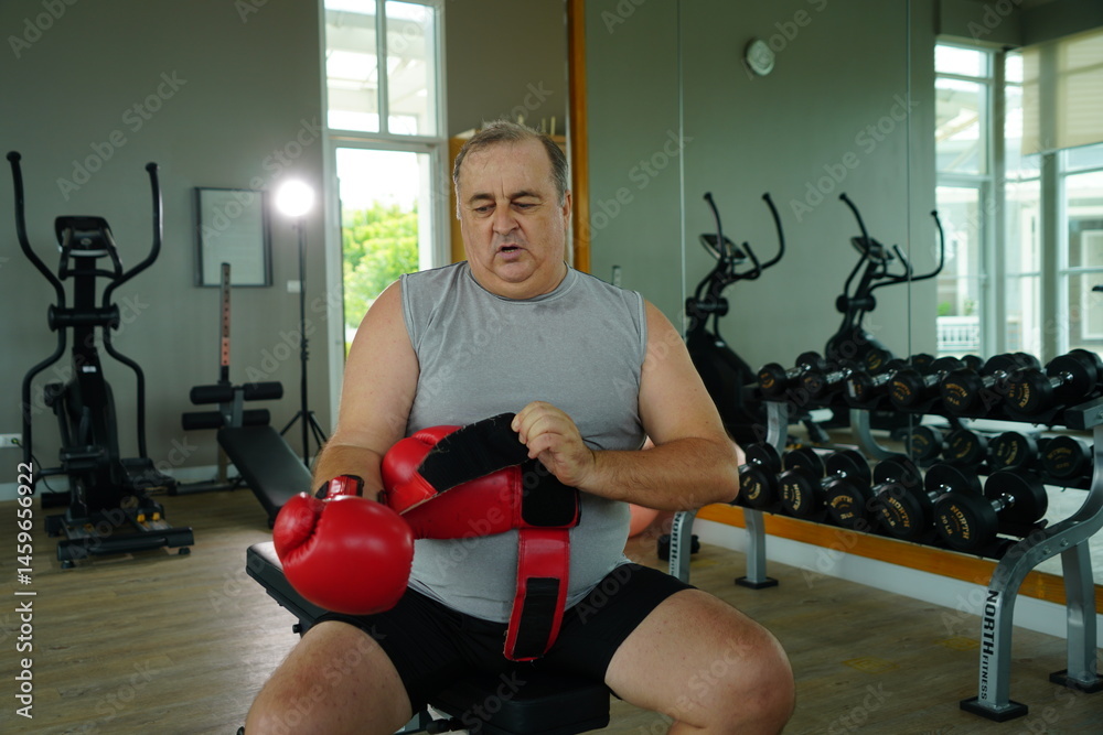 Elderly Caucasian man with a fat belly wearing gym clothes wearing red gloves practicing boxing and punching doing exercises for health and lose weight in a fitness centere.