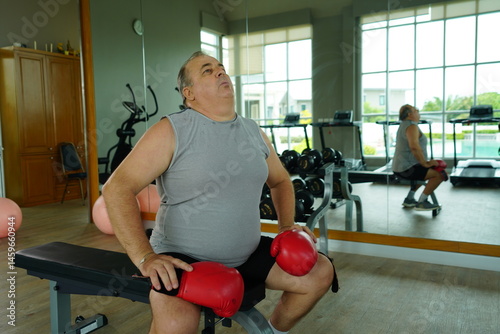 Obraz na płótnie Elderly Caucasian man with a fat belly wearing gym clothes wearing red gloves practicing boxing and punching doing exercises for health and lose weight in a fitness centere. w salonie