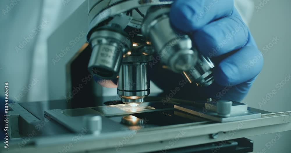 Close-up of biologist gloved hands using microscope in sterile laboratory. Vibrant blue gloves highlight precise scientific work against monochromatic lab backdrop