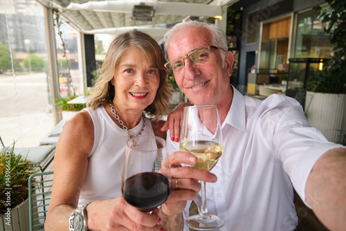 Obraz na płótnie Happy retired couple taking selfie while toasting wine glasses at outdoor restaurant w salonie