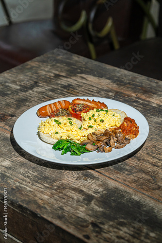 Big breakfast with bread toast, scrambled egg, sausages, roasted tomato, baked bean, sauted mushroom, and wilted spinach on white plate. Isolated by wooden table. Food Photography