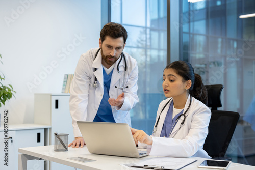Obraz na płótnie Two doctors collaborate in a modern office setting, focusing on a laptop while discussing medical information and data. The scene portrays professional teamwork. w salonie