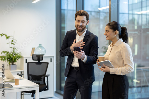 Obraz na płótnie A man and a woman smile while reviewing something on a smartphone in a modern office setting, holding a coffee and tablet. w salonie