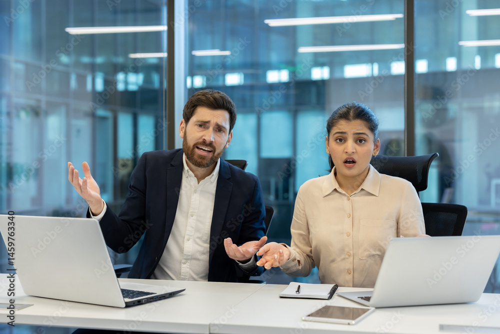 A shocked business team appears to be dealing with an unexpected issue while working at their office desk. They're using laptops.
