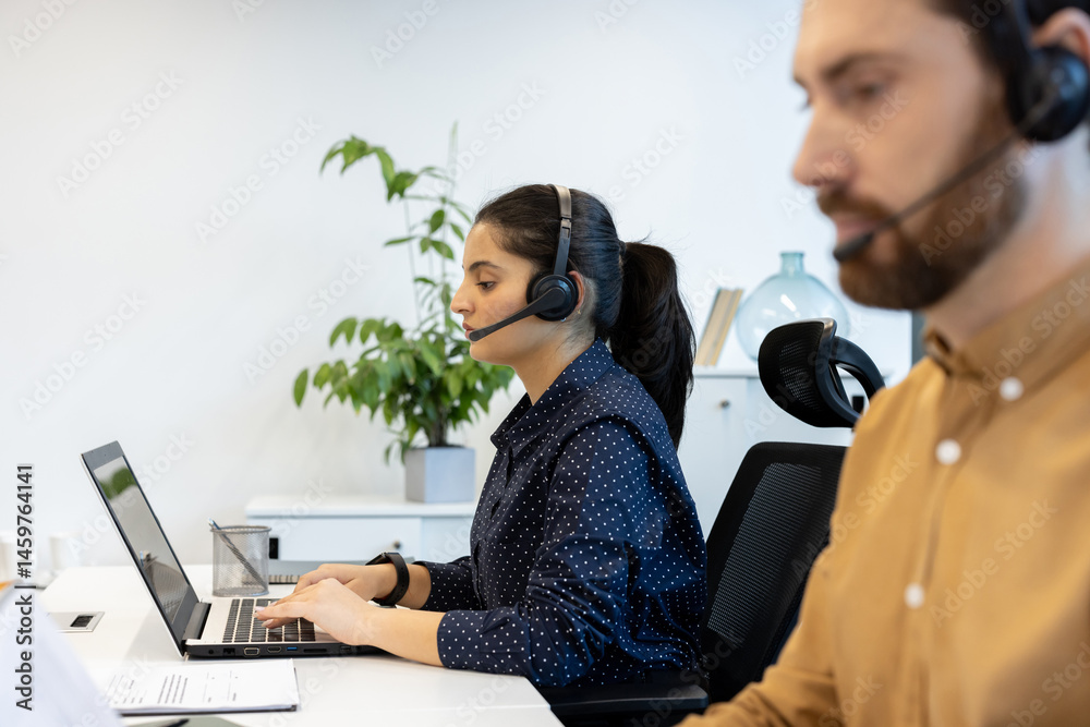 A call center employee with a headset is working on a laptop in a bright office environment. Another person is partially visible.