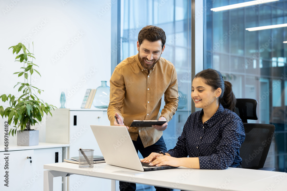 A cheerful colleague assists a female coworker as she works on a laptop in a bright modern office setting. Collaboration is key.
