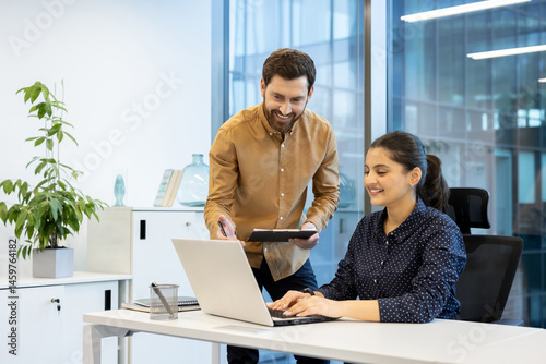 Obraz na płótnie A cheerful colleague assists a female coworker as she works on a laptop in a bright modern office setting. Collaboration is key. w salonie