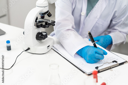 Closeup of asian scientist hand wearing blue glove writing research data on clipboard next to microscope in white laboratory, documenting analysis results during scientific experiment and observation