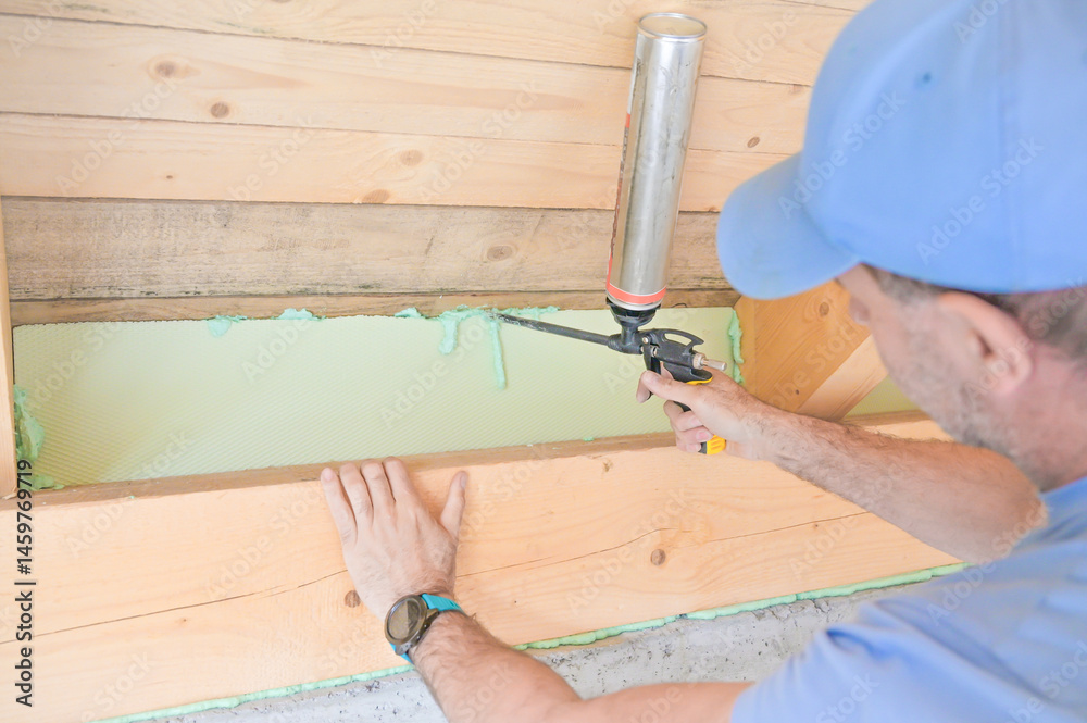A man uses a caulking gun to apply insulation along a wooden beam in a home renovation project, highlighting the importance of energy efficiency and construction skills