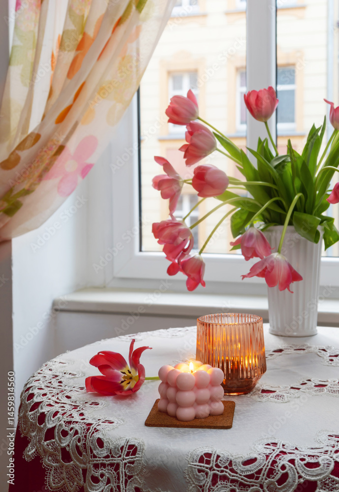 pink tulips in white vase with burning candles on background window
