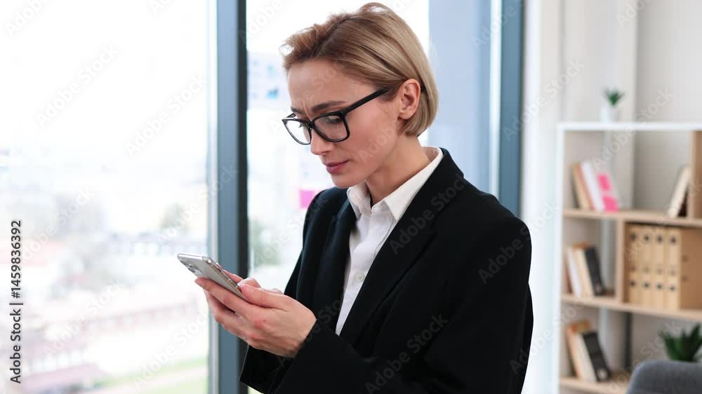 Confident middle-aged woman in formal attire standing in bright office holding smartphone smiling Confident middle-aged woman in formal attire standing in bright office holding smartphone smiling