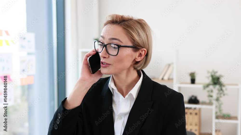 Blonde professional adult woman engaged in phone conversation standing in modern office wearing suit Blonde professional adult woman engaged in phone conversation standing in modern office wearing suit
