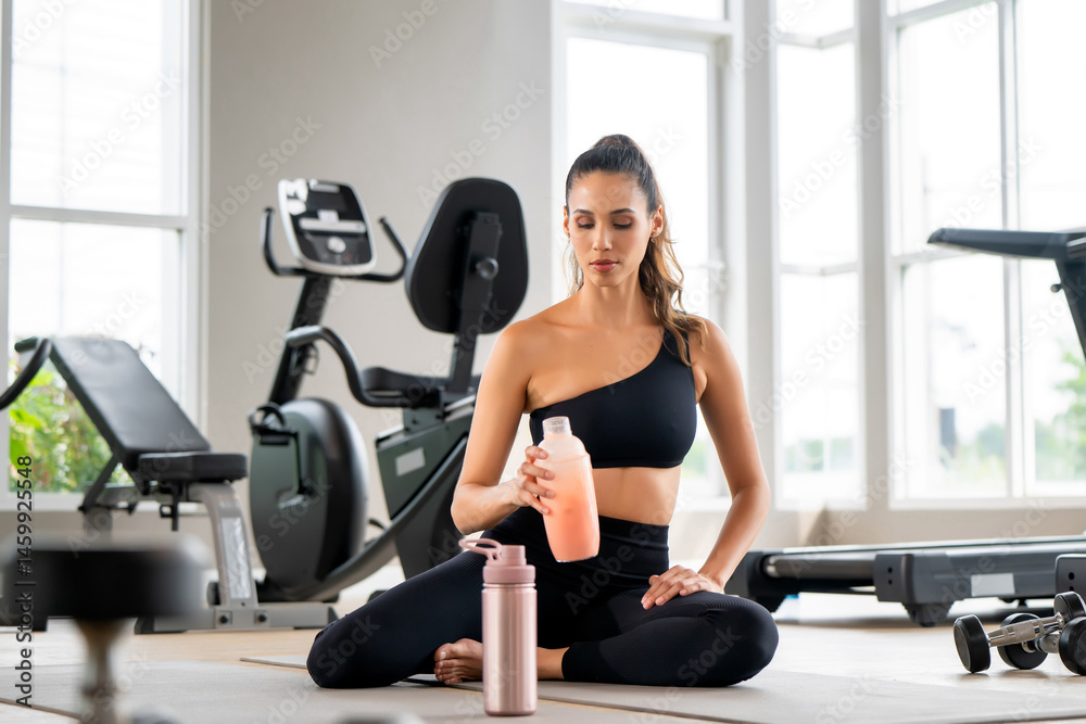 Fototapeta premium Confident woman in workout clothes sits on a yoga mat and holds a healthy strawberry protein smoothie after a workout in sport gym, showing strength, freshness and an active modern lifestyle.