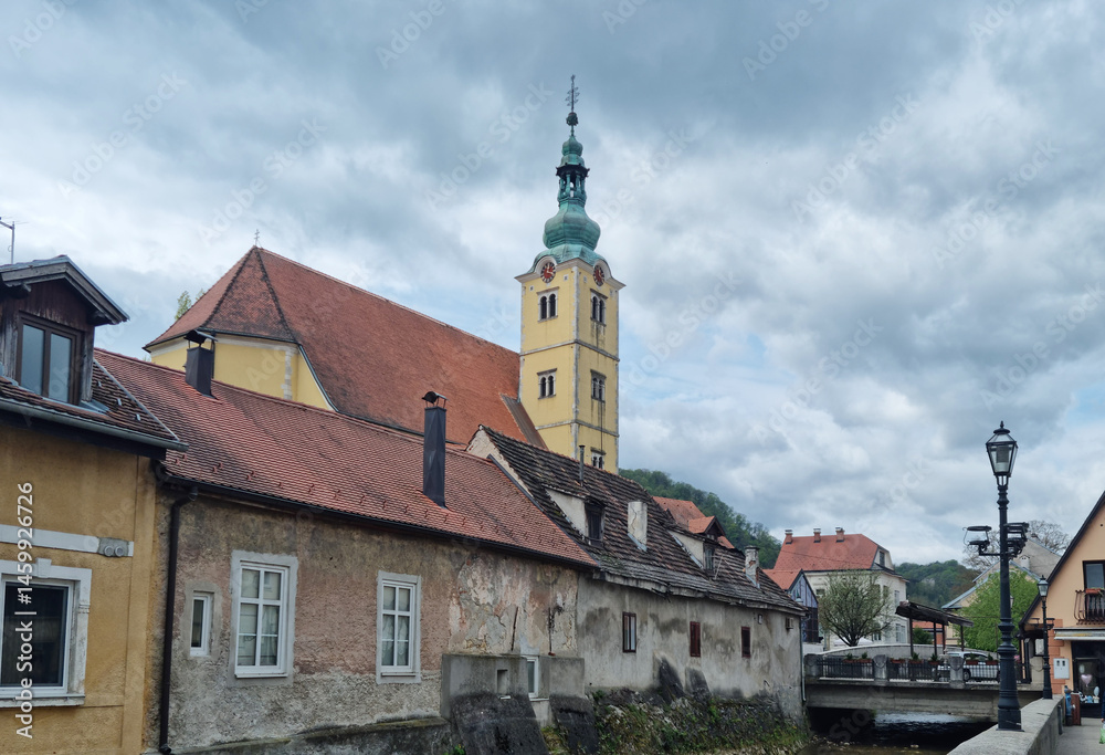 An overcast day in Samobor showcases the steeple of St. Anastasia Church rising above charming old buildings with red-tiled roofs along a waterway
