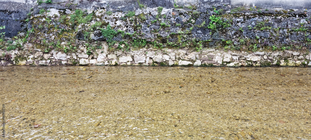 The shallow, clear water of the Gradna River flows gently along a moss-covered stone embankment in Samobor