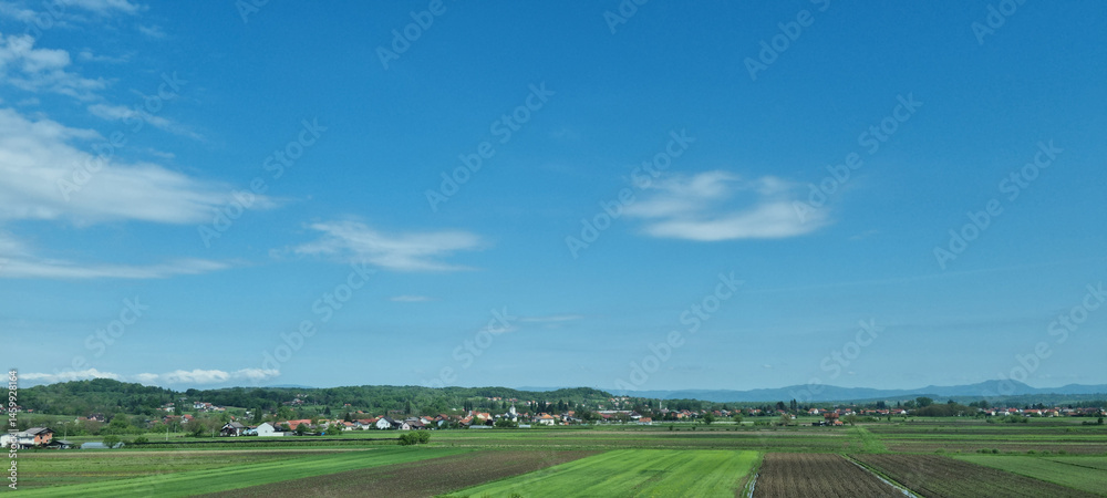 A vast agricultural landscape with fields and a distant town stretches under a bright blue sky with scattered clouds, viewed from the highway passing through Karlovac towards the north