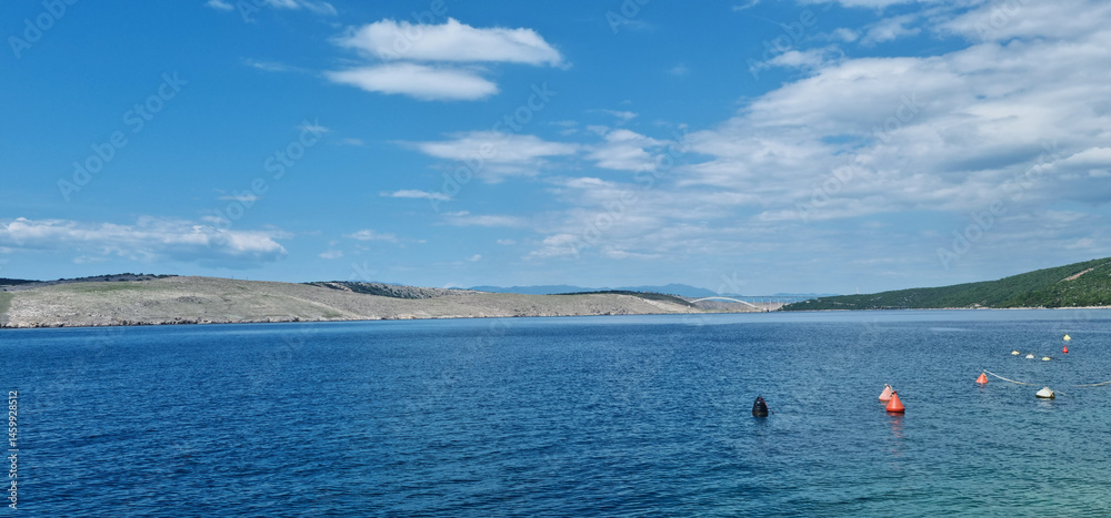 A scenic view from Jadranovo shows the calm, deep blue Adriatic Sea leading towards the distant arch of the Krk Bridge under a partly cloudy blue sky