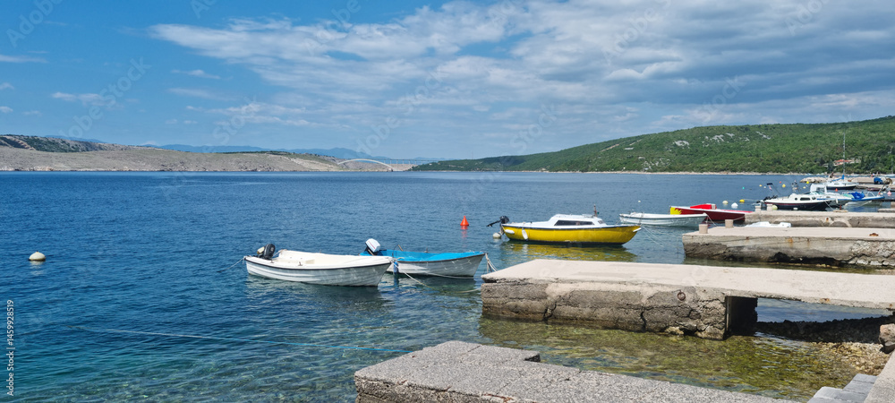 A picturesque harbor in Jadranovo features several small boats moored near concrete piers, with the Krk Bridge visible in the distance over the blue sea under a partly cloudy sky