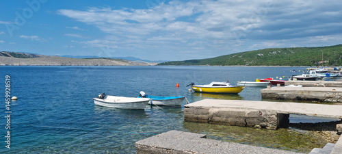 Obraz na płótnie A picturesque harbor in Jadranovo features several small boats moored near concrete piers, with the Krk Bridge visible in the distance over the blue sea under a partly cloudy sky w salonie