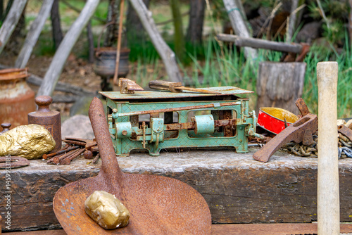 arrangement of old gold prospecting tools, including a rusty shovel with a fake gold nugget, a vintage scale, and various metal items placed on a weathered wooden table outdoors. The scene is styled t