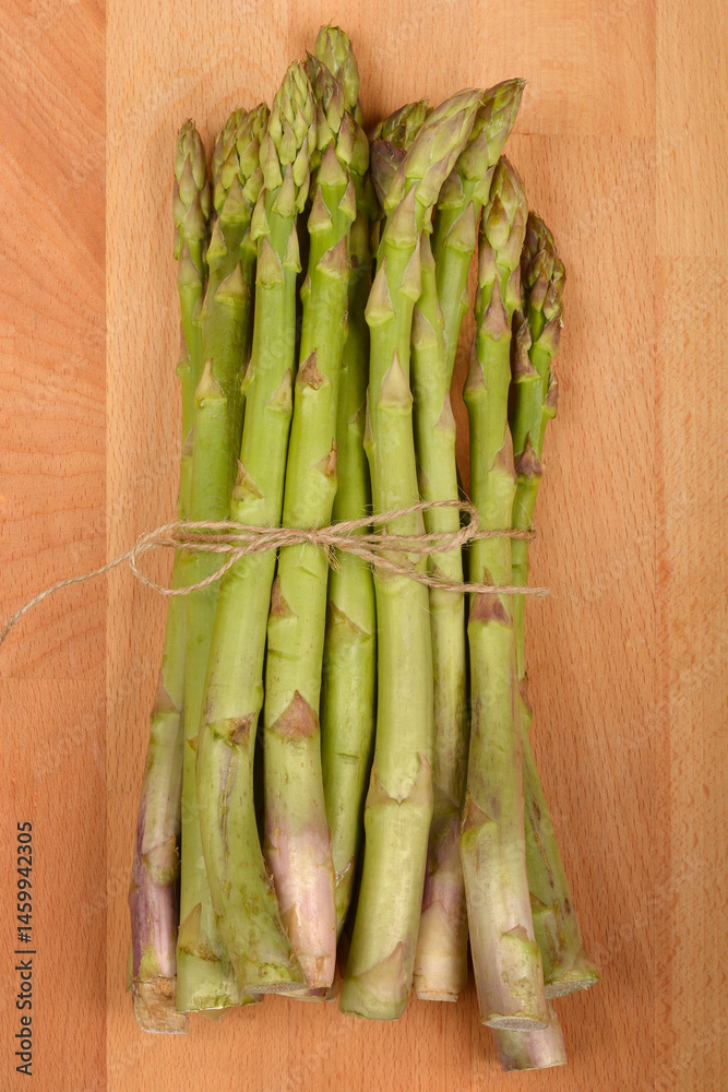A bundle of fresh green asparagus spears, tied together with twine, rests on a light brown wooden surface