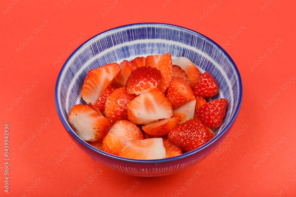A blue and white patterned bowl is filled with halved fresh red strawberries on a red background