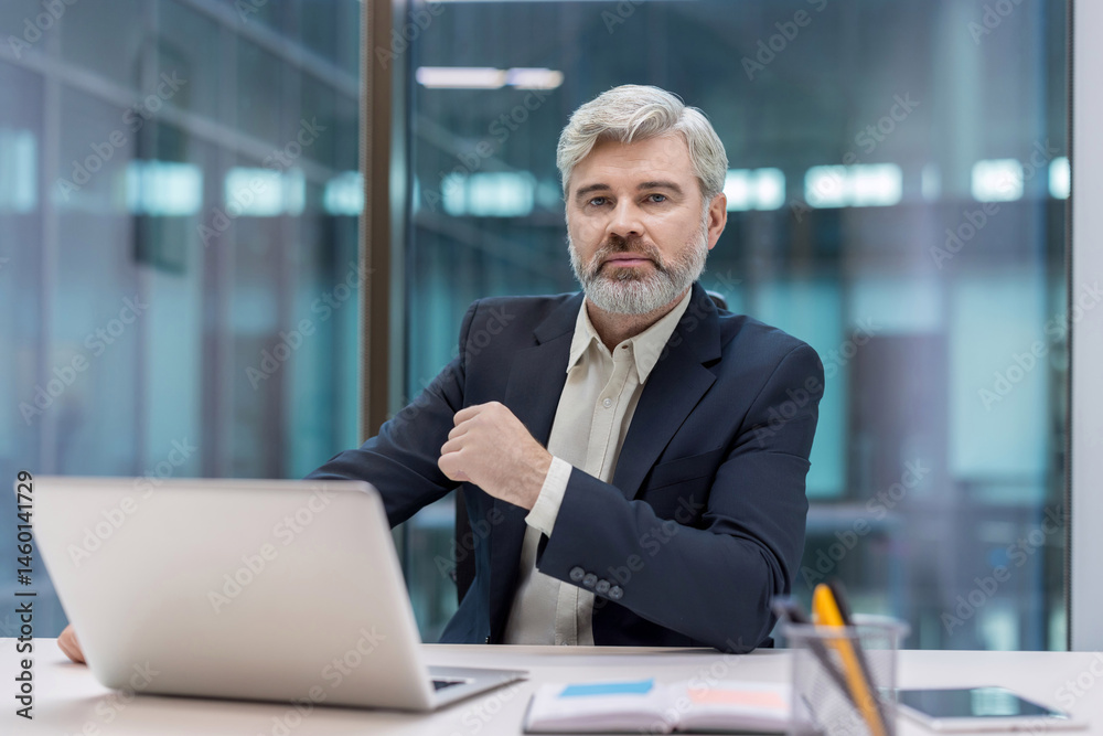 A distinguished-looking man with a gray beard sits at his desk, looking at the camera.