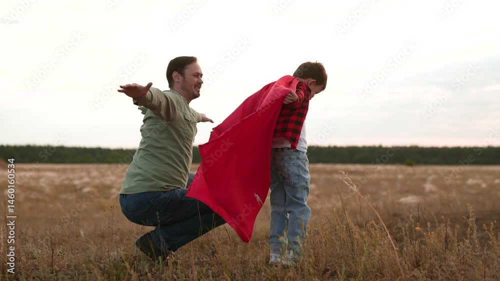 Father squats and son with red cape blowing by wind play superheroes . Father encourages son imitating airplane flight moving arms. Father with son feel lightness of freedom on field