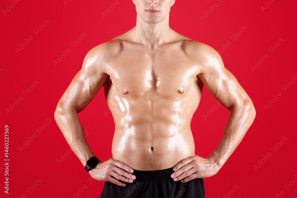 Fototapeta premium Handsome muscular young man with bare torso posing on red studio background. Serious millennial bodybuilder with shirtless chest and prominent muscles looking at camera. Sports and fitness concept
