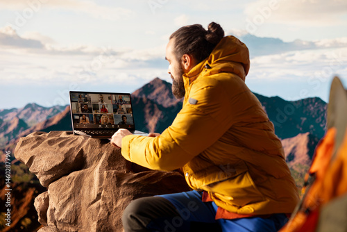 Young nature lover businessman meeting with colleagues on online video call, enjoying high speed internet and connectivity on top of the mountain. Male hiker entrepreneur with dependency.