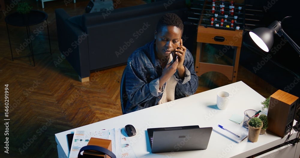 African american teleworker receiving phone call from job colleague while working from home on laptop. Freelancer speaking with coworker using smartphone while solving tasks, camera B