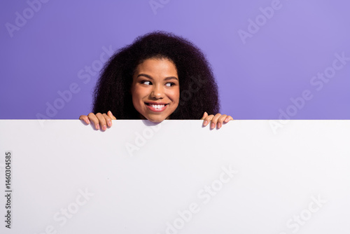 Obraz na płótnie Young woman peeking over a white surface with playful expression against a solid purple background w salonie