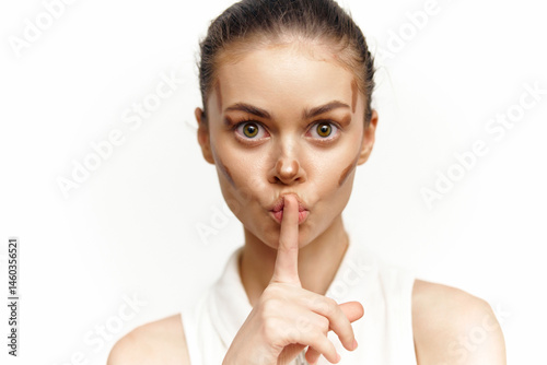 Obraz na płótnie A young woman with a serious expression holds a finger to her lips, wearing a white top, evoking a sense of mystery and intrigue. w salonie