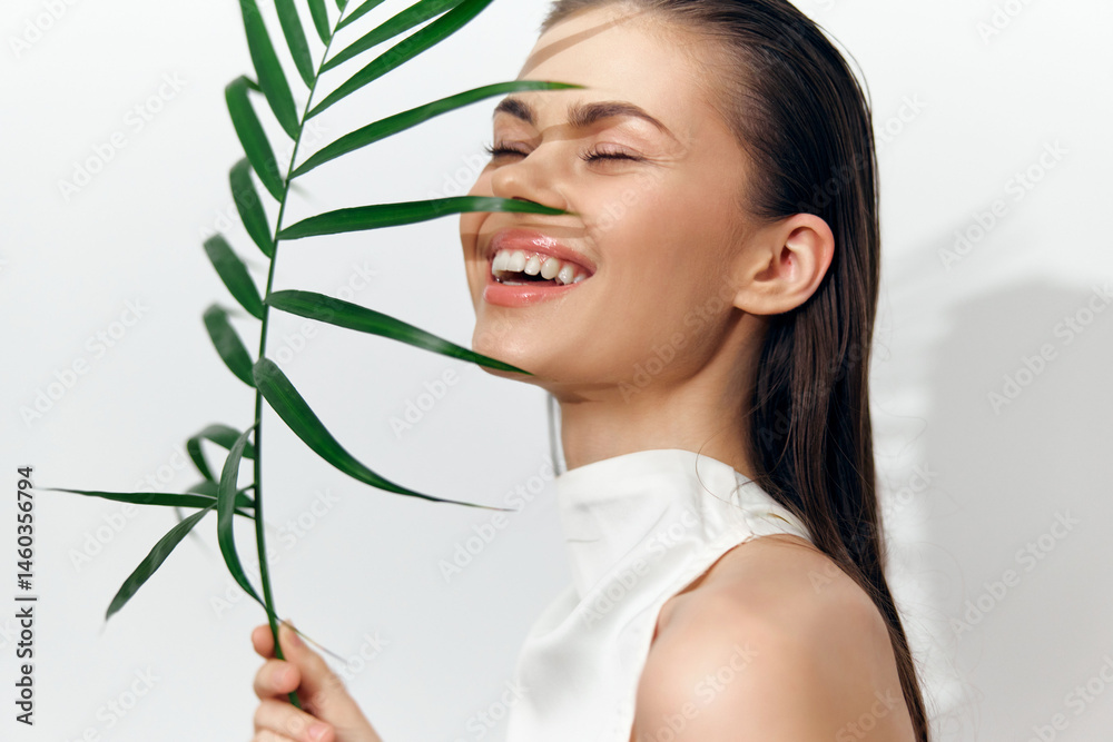 Joyful young woman smiling behind a green leaf, dressed in a stylish sleeveless outfit, exuding happiness and freshness in a minimalist setting.