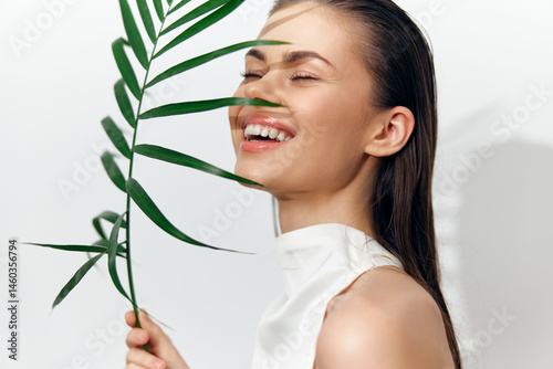 Obraz na płótnie Joyful young woman smiling behind a green leaf, dressed in a stylish sleeveless outfit, exuding happiness and freshness in a minimalist setting. w salonie