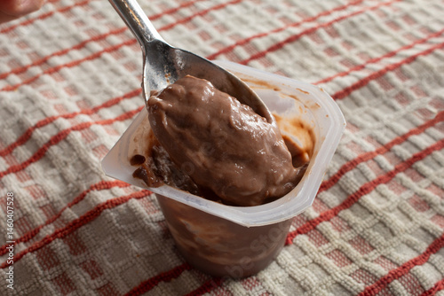 A view of a package cup of chocolate pudding. A spoon scoops a spoonful.