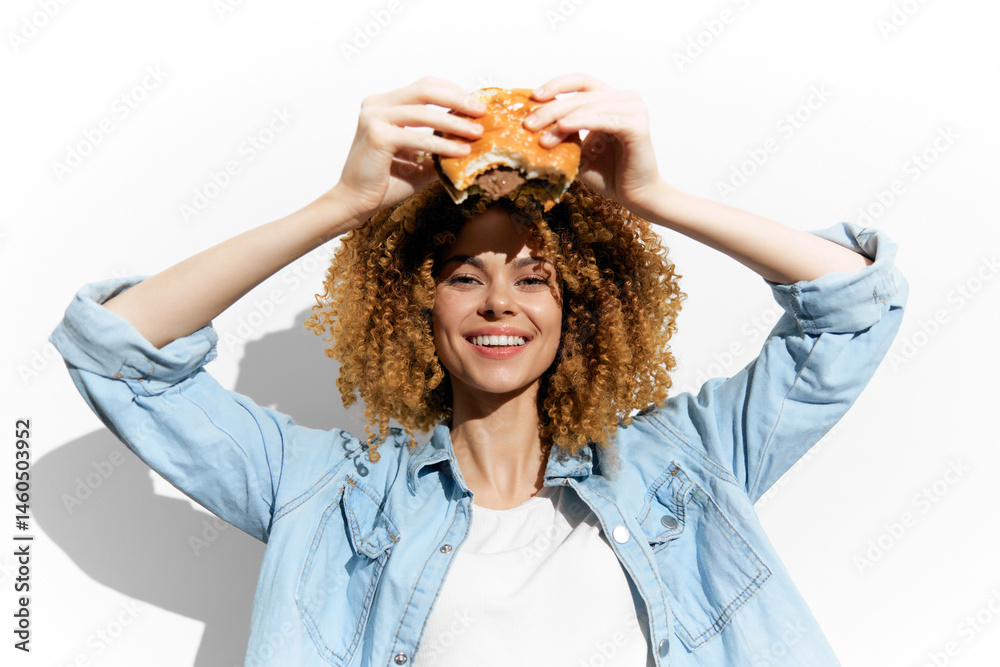 Cheerful woman with curly hair holding a burger, showcasing a fun food concept in a bright, minimalist setting Emphasizing joy and indulgence in fast food
